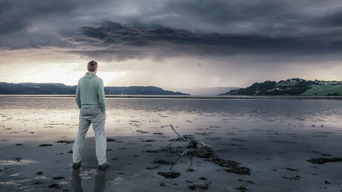 Man looking out over a gray lake with rain clouds appearing on horizon