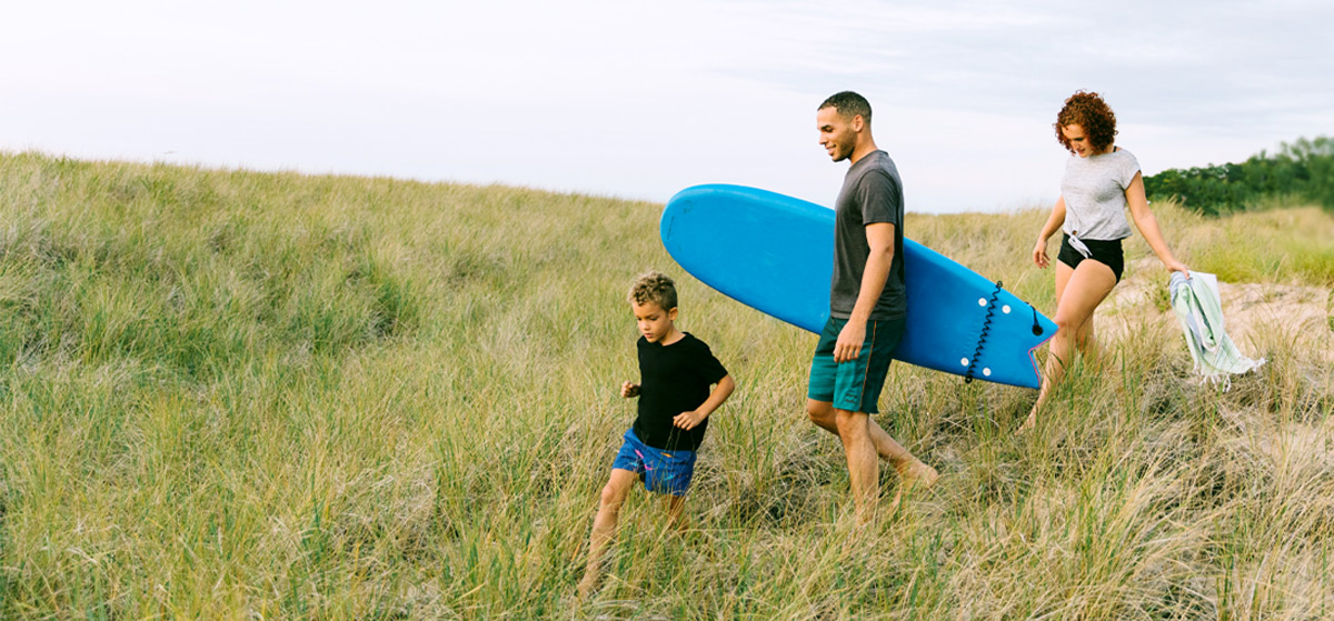 Photo of a husband, wife, and their young son walking down to the beach while carrying a boogie board